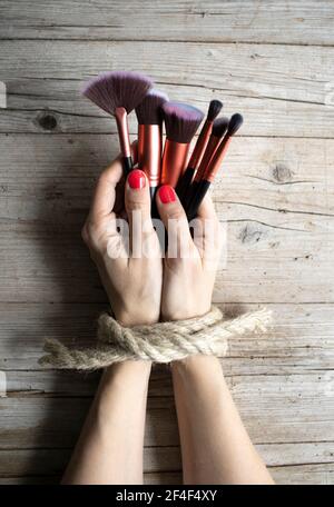Hands of young woman with red fingernails are tied and lie on light wooden background, she holds makeup brushes, concept about addiction, beauty craze Stock Photo