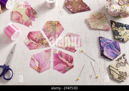 Pentagonal pieces of fabric lying in the shape of a flower, stacks of multi-colored pieces of fabric, sewing accessories on a white surface