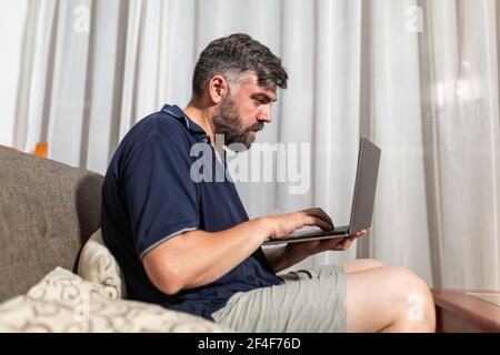 bearded man on sofa uses laptop in dining room overnight Stock Photo