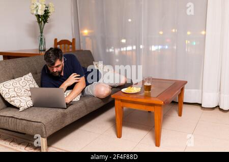 bearded man on sofa uses laptop in dining room overnight Stock Photo