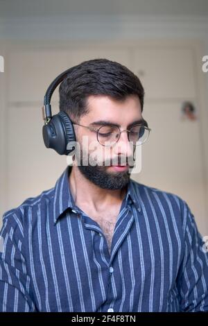 A man with a beard and a striped shirt listening to music with headphones in his room Stock Photo