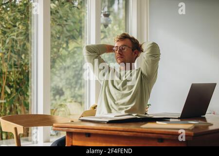 Man sitting at table with eyes closed and hands behind head at home. Man taking break from work. Stock Photo
