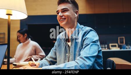 Smiling male student with earphones studying in college library. Happy young man in university looking away and smiling. Stock Photo