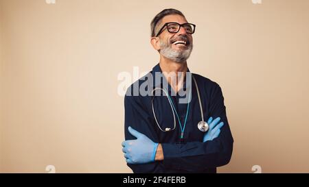 Portrait of a cheerful general practitioner with stethoscope wearing protective hand gloves. Doctor standing with his arms crossed looking away and sm Stock Photo