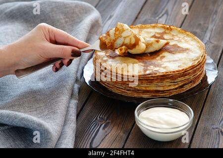 A young woman picks up a pancake with a fork. Sour cream for pancakes on the table, breakfast for the whole family. Maslenitsa, the holiday of the end Stock Photo