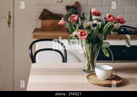 Spring breakfast still life with cup of coffee. Red tulips, white anemone flowers and eucalyptus branches. Glass vase on wooden table. Blurred kitchen Stock Photo