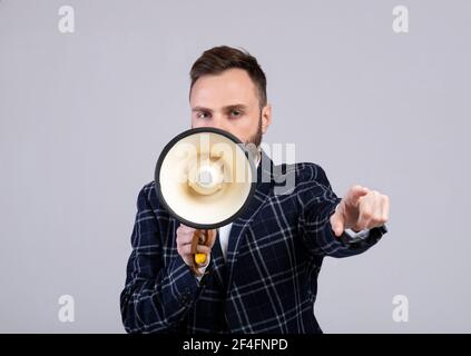 Determined young man in formal wear shouting into megaphone and pointing at you on grey studio background Stock Photo