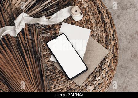 Tropical summer mockups. Black cell phone with empty screen, blank greeting card, envelope and ribbon placed on rattan straw pouff. Dry palm leaves Stock Photo