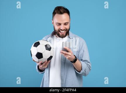 Happy young guy with soccer ball using smartphone, winning sports bet, rooting for his favorite team on blue background Stock Photo