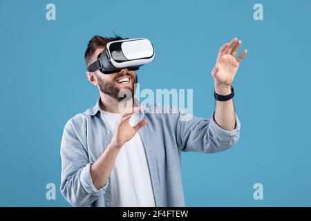 Happy bearded young man using VR headset to explore augmented reality on blue studio background Stock Photo