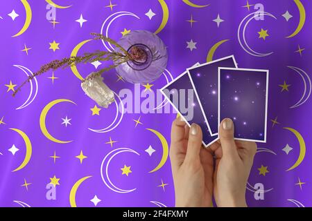 Hands of young woman with golden nail polish holding three tarot or oracle cards, on a purple stars and crescent pattern table cloth Stock Photo