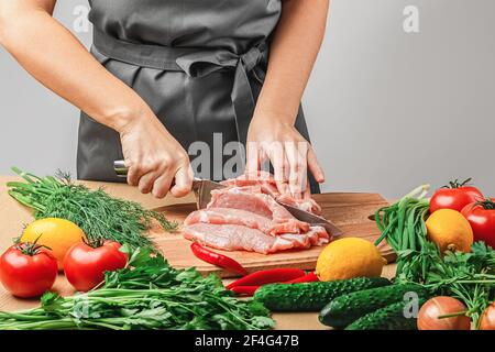 A woman in a gray apron cuts a piece of meat with a knife on a wooden board. There are onions, tomatoes, dill, parsley, tomatoes, red hot peppers and Stock Photo