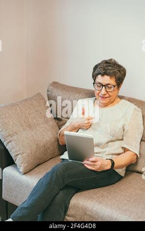 Attractive elegant senior woman sitting on the couch and using a tablet computer at home. Stock Photo