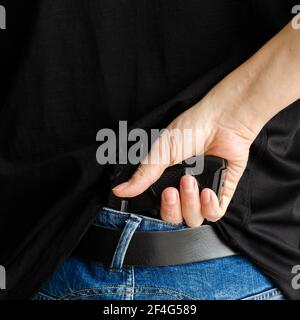 Hided handgun under the denim belt. a man in jeans and a t-shirt holds a gun in his hand from the back Stock Photo