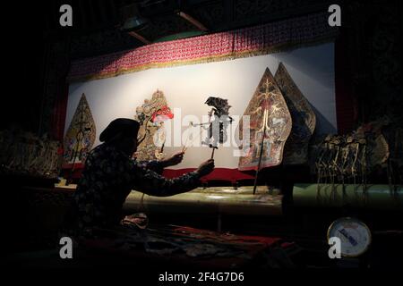 Puppet master (dalang) performs the wayang kulit performance 'The Death of Kumbakarna' in the Sonobudoyo Museum in Yogyakarta, Central Java, Indonesia. The Kumbhakarna puppet are seen on the screen at the left while the allegorical figures of the Gunungan representing both the Javanese Tree of Life and the Cosmic Mountain (Mount Meru) are seen on each side of the screen. Traditional puppet shadow theatre known as wayang kulit is widespread on the islands of Java and Bali in Indonesia. Each wayang kulit performance continues about eight hours through the night without intermissions and only one Stock Photo