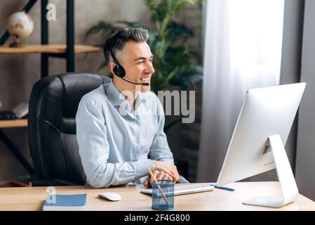 A joyful caucasian senior business leader, manager, sits at a table, at the office, in a stylish shirt, communicates via a video call with employees or friends, using a headset and computer, smiling Stock Photo