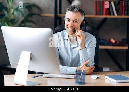 Caucasian confident successful businessman, coach, sitting at a table in the office, wearing headphones and formal shirt, uses computer, works remotely , looking at the camera with friendly smiles Stock Photo