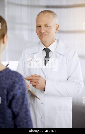 Elderly confident doctor consults woman-patient in sunny clinic. Perfect medical service in medicine Stock Photo