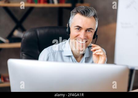Portrait of a confident successful senior grey-haired Caucasian office manager, businessman or leader, worker of call center, wearing headset, sits at workplace looks at camera and friendly smiling Stock Photo