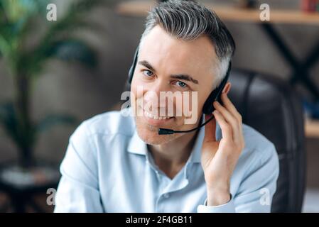 Portrait of a smiling confident middle aged caucasian office worker working remotely, office manager, businessman or leader, wearing headset, sits at workplace looks at camera and friendly smiling Stock Photo