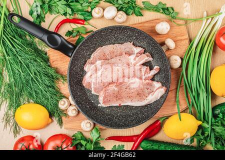 Top view of slices of chopped raw pork with spices on a black frying pan and a wooden board. On the table are lemons, tomatoes, hot peppers, mushrooms