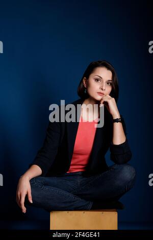 Portrait of young brunette woman wearing a coral tank top, black blazer and blue jeans on dark blue background. Stock Photo