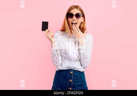 Happy worried amazed young woman holding credit card on pink background Stock Photo