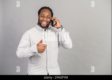 Black happy man talking on the phone over isolated grey background, showing the ok sign, thumb up with fingers, excellent sign, everything is under control, confident man settled conflict, made a deal Stock Photo