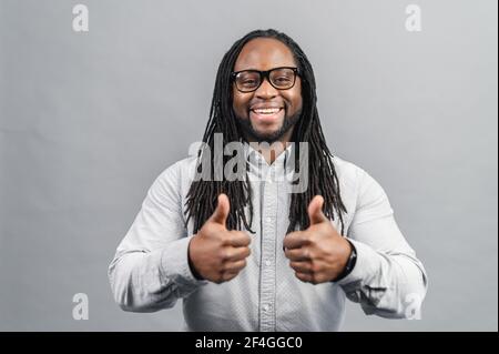 Positive African man with long dreadlocks, standing on a grey background, showing that everything is fine, looking at camera, showing thumbs up, doing approval gesture with fingers and both hands Stock Photo