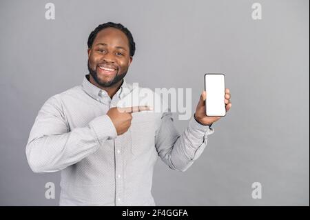 Portrait of cheerful, positive, attractive African american guy with stubble in casual shirt, having smart phone with white screen in hand, pointing with forefinger to product, on grey background Stock Photo