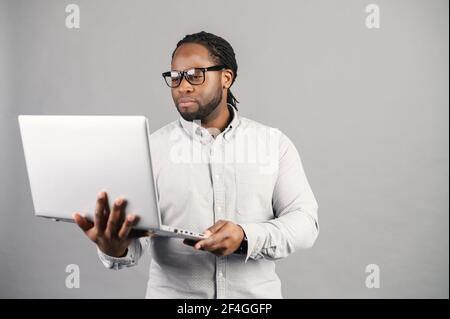 Concept of confident business expert. Serious young handsome black man with elegant glasses in casual shirt, holding laptop, looking at screen, standing against grey background, no time for break Stock Photo