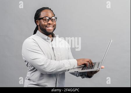 Smiling african man using laptop, standing at grey backdrop, checking home finance, happy man looking at invoice, looking at camera, connection with bank, checking bill using online application Stock Photo