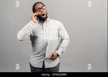 Young latin business man talking on the phone over isolated grey background stressed with laptop in the hand, shocked with shame and surprise face, angry and frustrated. Fear and upset for mistake. Stock Photo