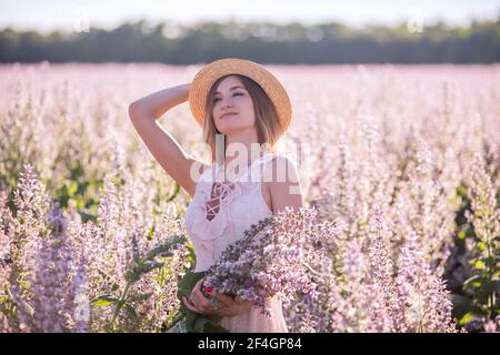 Young blonde woman in straw hat looks into the distance against the background of blooming field pink sage. Close-up portrait of beautiful girl holdin Stock Photo