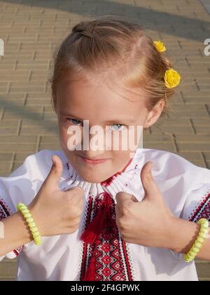Caucasian blonde little girl in festive Ukrainian embroidered shirt gestures all the best, summer time, sunny weather, Independence Day Stock Photo