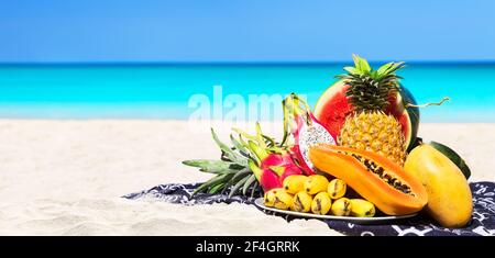 Panorama of fresh different tropical fruits placed on the beach with blue sky and sea background. Summer time beach concept of healthy eating. Stock Photo