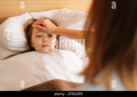 Close-up view of young mother measures temperature touches forehead of sick daughter lying in bed at home.  Stock Photo