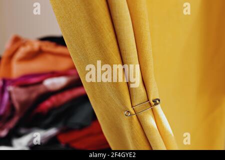 selective focus photo of a yellow curtain with an iron pin, with blurred multicolored textile clothes on the background Stock Photo