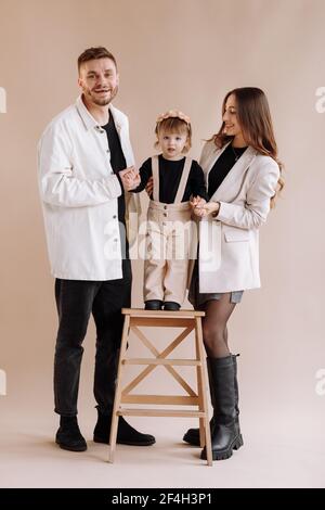 Portrait of a happy young family, with a little adorable daughter. parents holding a baby's hands and hug her. child standing on a wooden ladder with Stock Photo