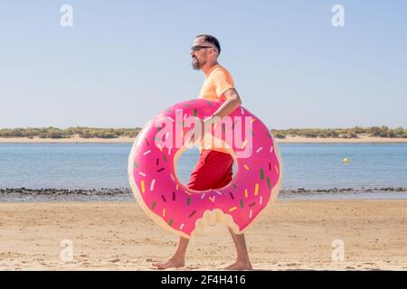 Young hipster man with a beard, swimming trunks and sunglasses carries a pink inflatable float on the beach. leisure on a sunny day on vacation Stock Photo