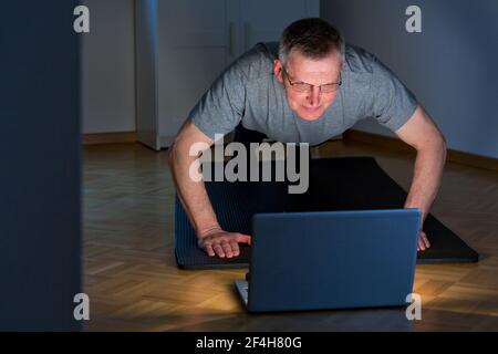 Mature or senior man doing push ups in a dark room at home in front of laptop - focus on the face Stock Photo