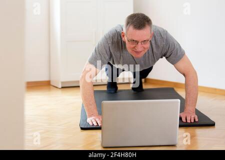 Mature or senior man doing push ups at home in front of laptop - focus on the face Stock Photo