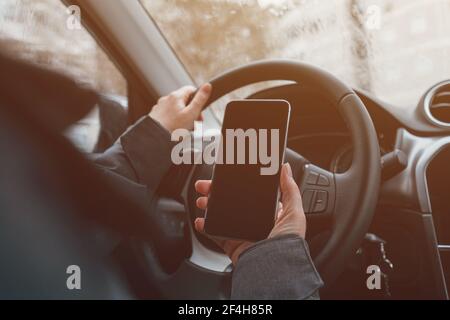 Looking at mobile phone screen while driving, woman using smartphone and steering the car Stock Photo