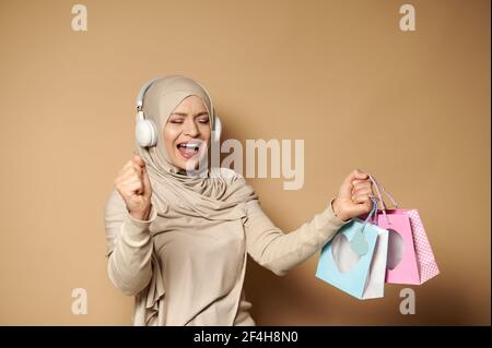 Muslim woman in hijab with headphones holding colored paper bags in her hands enjoying the upcoming religious holidays. Eid mubarak Said. Stock Photo