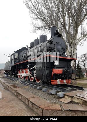 An old black steam locomotive on rails and a concrete platform in the middle of a city square. Stock Photo