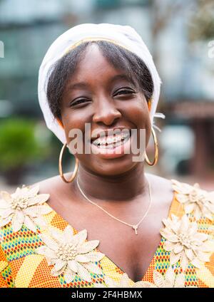 Young positive plump African American lady in elegant blue dress and wristwatch conversing on cellphone while sitting near stone wall and looking away Stock Photo