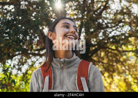 Happy optimistic young female hiker in activewear with backpack enjoying journey in green forest in sunny day Stock Photo