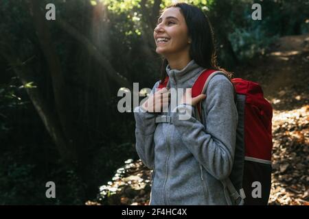 Happy optimistic young female hiker in activewear with backpack enjoying journey in green forest in sunny day Stock Photo