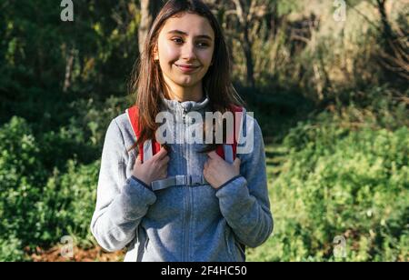 Happy optimistic young female hiker in activewear with backpack enjoying journey in green forest in sunny day Stock Photo
