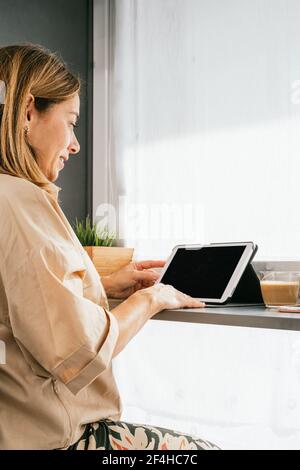 Side view of mature female sitting at counter in kitchen and browsing tablet in morning Stock Photo